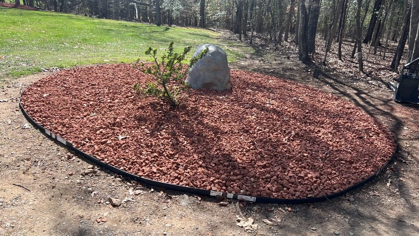 Round bed of red mulch with a boulder and small shrub on a wooded lot